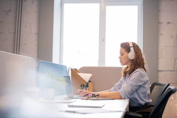 ernst beschäftigt attraktive junge Frau mit lockigem Haar trägt weiße Kopfhörer sitzt am Tisch und tippt auf Laptop im Büro - Foto, Bild
