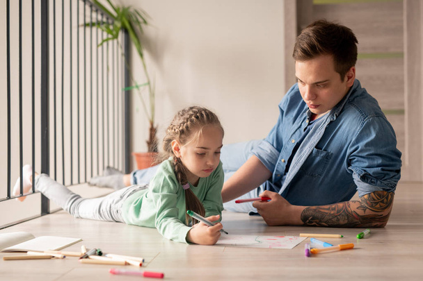 ernster Hipster junger Vater im Jeanshemd, der auf dem Boden liegt und der Tochter Ratschläge gibt, kleines Mädchen zeichnet Bild mit Vater zu Hause - Foto, Bild