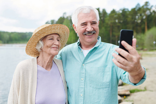 Ευτυχισμένο ρομαντικό ζευγάρι ηλικιωμένων στο ανεπίσημο ντύσιμο φτιάχνοντας selfie σε φυσικό περιβάλλον την καλοκαιρινή μέρα - Φωτογραφία, εικόνα
