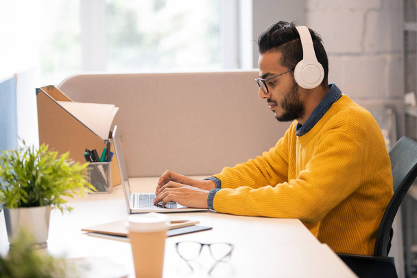 nachdenklicher junger arabischer Vermarkter mit Bart sitzt am Schreibtisch und hört Musik in drahtlosen Kopfhörern, während er mit Laptop arbeitet - Foto, Bild