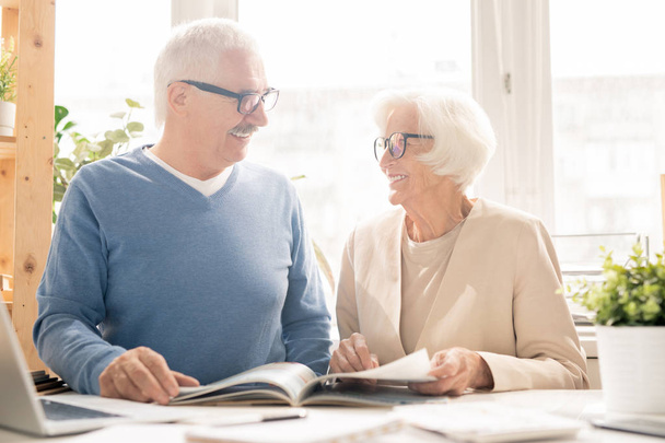 Akıllı casualwear mutlu yaşlı çift masa başında otururken birbirlerine bakarak ve dergi ile bakıyor - Fotoğraf, Görsel