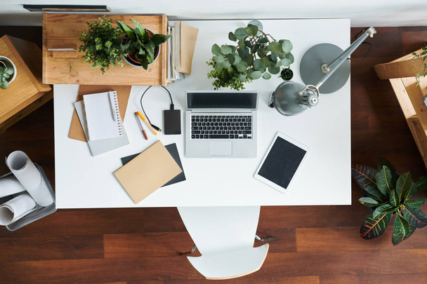 Офисные вещи на столе - тачпад, ноутбук с зарядным устройством, тетради, ручки, ноутбук, лампа и зеленые растения в цветочных горшках
 - Фото, изображение