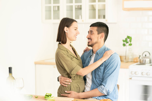 Молодий чоловік змішаної раси обіймає свою щасливу дружину на кухні за столом зі свіжими овочами та пляшкою вина
 - Фото, зображення