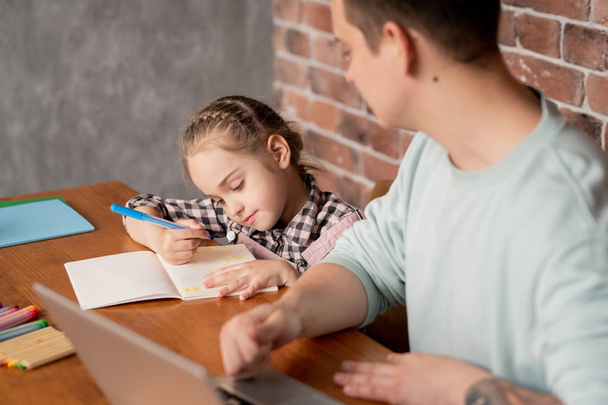 Content cute little daughter with braid sitting at table and showing drawing in notebook to father working with laptop at home - 写真・画像