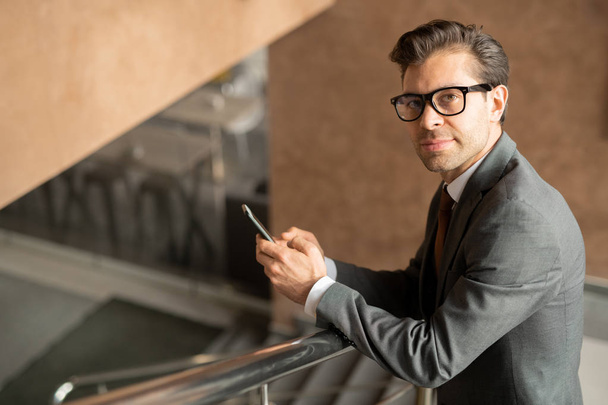 Young confident male business agent looking at you while standing in front of camera by railings and scrolling in smartphone - Photo, Image