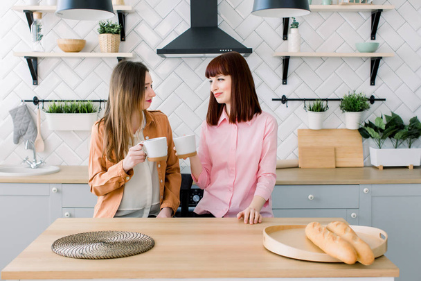 Two Female Caucasian Friends sipping coffee in kitchen - Foto, afbeelding