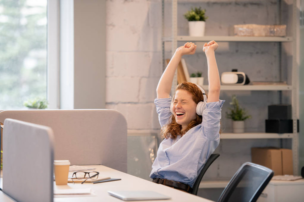 Веселая молодая женщина-менеджер с рыжими волосами сидит за столом в офисе и поднимает руки, радуясь успешной реализации проекта
 - Фото, изображение