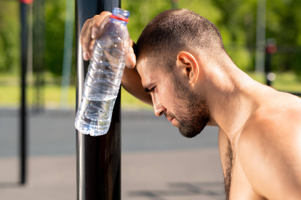 Atleta stanco con bottiglia d'acqua appoggiata agli impianti sportivi mentre riposa dopo un duro allenamento all'aria aperta
 - Foto, immagini