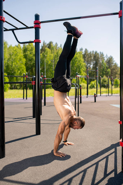 Topless young sportsman in black leggins, shorts and sneakers standing on hands over sportsground outdoors - Fotoğraf, Görsel