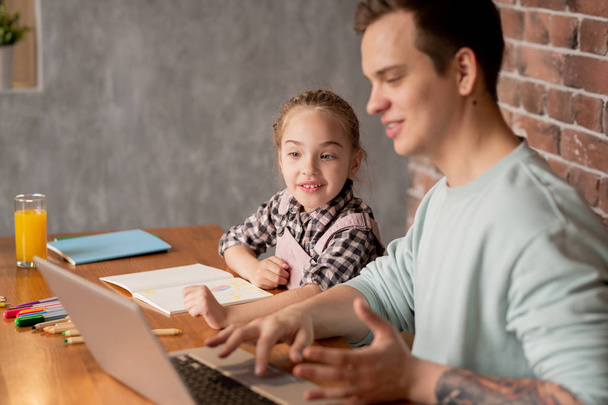 Menina curiosa positiva com trança sentada à mesa e usando laptop com o pai mostrando aplicativo de aprendizagem enquanto eles fazem a tarefa de casa criativa juntos
 - Foto, Imagem