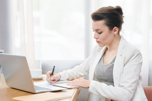 Serious concentrated businesswoman in white jacket sitting at table in restaurant and making notes in notebook while composing business plan - Foto, Imagen