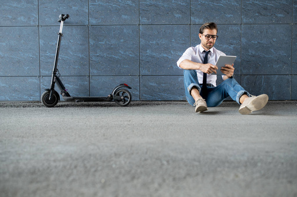 Jeune homme mobile élégant avec gadget moderne naviguant dans le net tout en étant assis sur le sol par un mur de construction
 - Photo, image