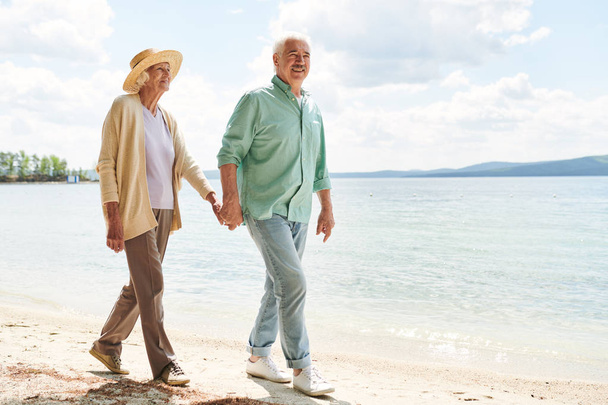 W wieku dorywczo mężczyzna i kobieta trzymający ręce podczas chłodzenia na plaży wzdłuż wybrzeża w gorącym letnim dniu - Zdjęcie, obraz