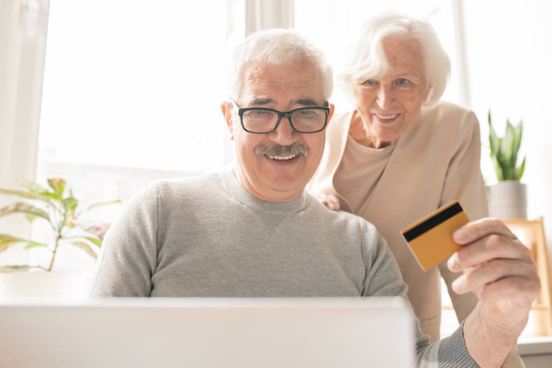 Zeitgenössischer Senior mit Plastikkarte sitzt vor dem Laptop, während er online einkauft und Bestellungen bezahlt - Foto, Bild