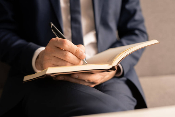 Χέρι του πολυάσχολο νεαρό αρσενικό επιχειρηματία κρατώντας στυλό πάνω από τη σελίδα του ανοιχτού σημειωματαρίου, ενώ κάνουν επιχειρηματικές σημειώσεις - Φωτογραφία, εικόνα