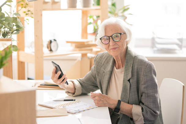 Σύγχρονη και κινητή ανώτερη γυναίκα σε τυποφόρα και γυαλιά που κάθονται από το χώρο εργασίας στο γραφείο μπροστά από την κάμερα - Φωτογραφία, εικόνα