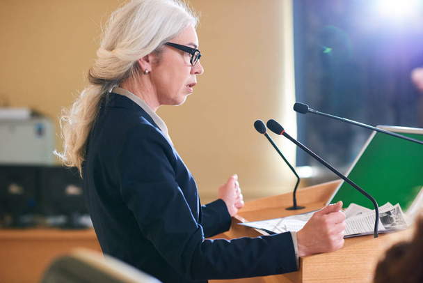 Maturo elegante delegata donna in giacca e cravatta che parla in microfono in piedi accanto al tribuno nella sala conferenze al vertice
 - Foto, immagini