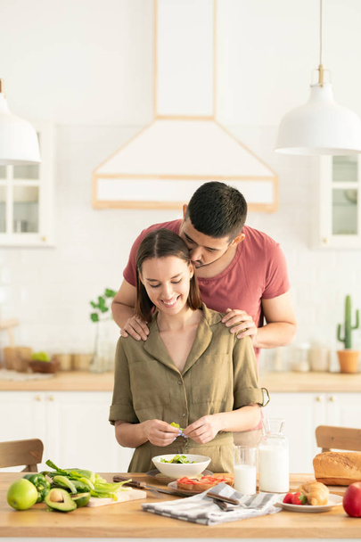 Молодий веселий чоловік дає дружині поцілунок на її голову, коли вона готує овочевий салат на сніданок
 - Фото, зображення