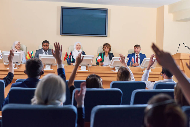 Público em poltronas levantando as mãos t oask perguntas para delegados interculturais em negócios ou evento político
 - Foto, Imagem