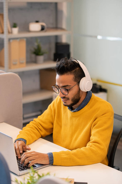 Serious young middle-eastern business intern with beard and mustache sitting at desk in office and using laptop while answering customer online - 写真・画像