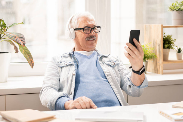 Современный зрелый мобильный мужчина в повседневной одежде и наушниках, смотрящий видео в смартфоне, сидя в офисе
 - Фото, изображение
