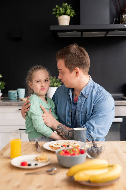 Picky daughter sitting on fathers knees and grimacing while refusing to eat breakfast made by father - Foto, imagen
