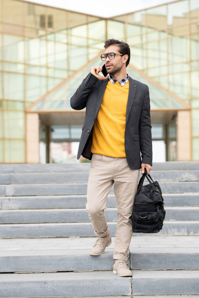 junger Geschäftsreisender mit Gepäcktasche bewegt sich die Treppe hinunter, während er im städtischen Umfeld mit dem Smartphone spricht - Foto, Bild