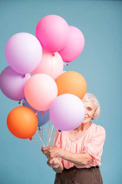Happy senior woman peeking out of bunch of colorful balloons in isolation - 写真・画像