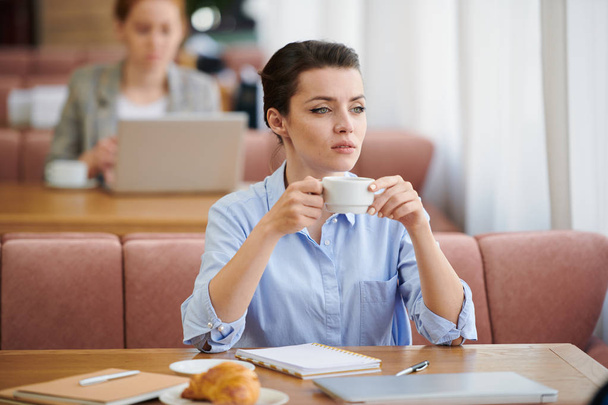 Serious purposeful young brunette businesswoman in blue shirt sitting at table and drinking coffee while having break in cafe - Foto, imagen