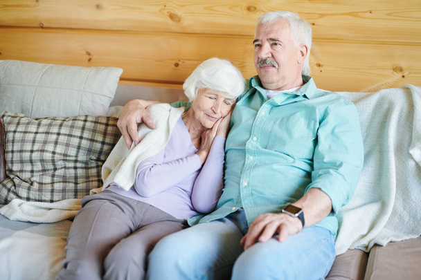Hombre mayor en ropa de casualwear sentado en el sofá y viendo el programa de televisión mientras su esposa siesta a su lado en la noche
 - Foto, imagen