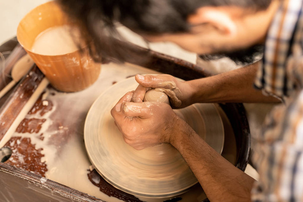Mãos de jovem mestre no centro da roda de cerâmica rotativa durante o trabalho sobre vaso novo ou outro item de barro
 - Foto, Imagem