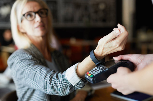Mulher loira moderna mantendo seu pulso sobre a máquina de pagamento eletrônico enquanto paga a ordem através de smartwatch no café
 - Foto, Imagem