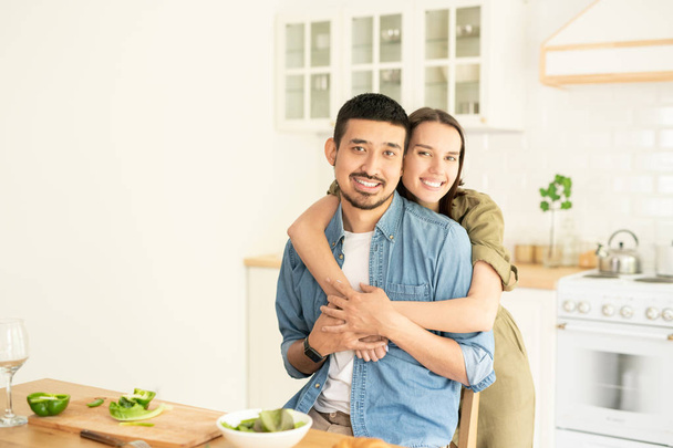 Young woman giving her husband hug while both looking at you in front of kitchen table while preparing breakfast - Photo, Image