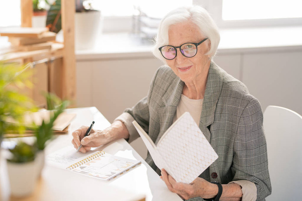 Feliz mujer de negocios de pelo blanco senior con libro abierto haciendo notas en el cuaderno mientras lee por escritorio
 - Foto, imagen