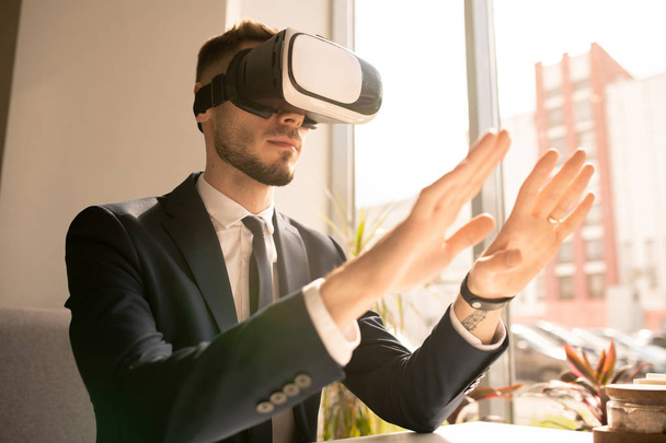 Współczesny młody biznesmen w VR Headset dotykając wirtualnego obiektu lub wyświetlacza siedząc w kawiarni przez okno - Zdjęcie, obraz