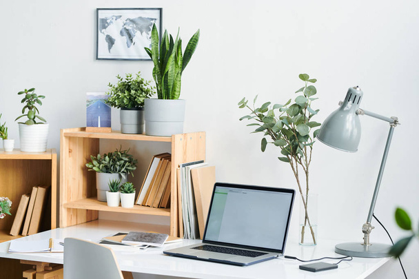 Candeeiro de mesa, laptop, cadernos e livros em mesa e prateleiras com plantas domésticas verdes em vasos de flores dentro do escritório
 - Foto, Imagem