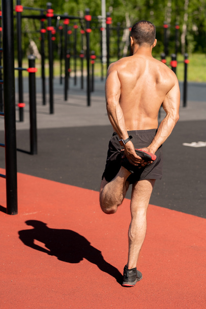 Vista trasera del joven deportista sin camisa sosteniendo su pie izquierdo en la mano mientras se calienta antes de entrenar en instalaciones deportivas
 - Foto, Imagen