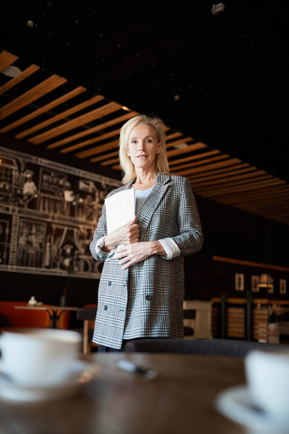 Blonde femme d'affaires mature avec des documents par poitrine debout à côté d'une des tables dans un restaurant moderne et vous regardant
 - Photo, image