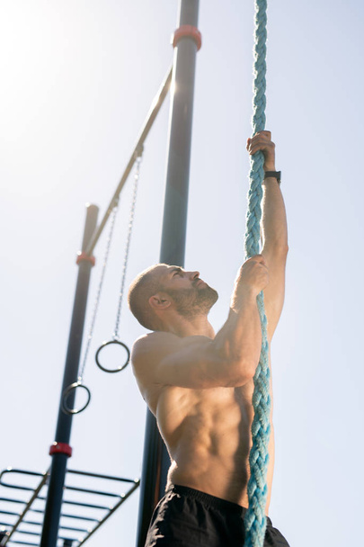 Jovem atleta muscular olhando para cima enquanto sobe corda grossa pendurada em um dos bares esportivos ao ar livre
 - Foto, Imagem