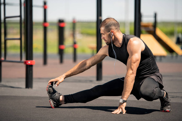 Jovem atleta ativo em traje preto esticando seu braço direito e perna enquanto se exercita no sportsground ao ar livre
 - Foto, Imagem