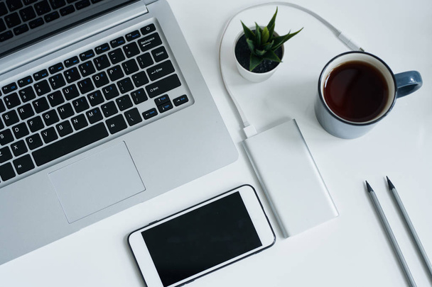 Вид клавіатури ноутбука, смартфон, кухоль з кавою або чаєм, зелена рослина в маленькому горщику, два олівці і зарядний пристрій на столі
 - Фото, зображення