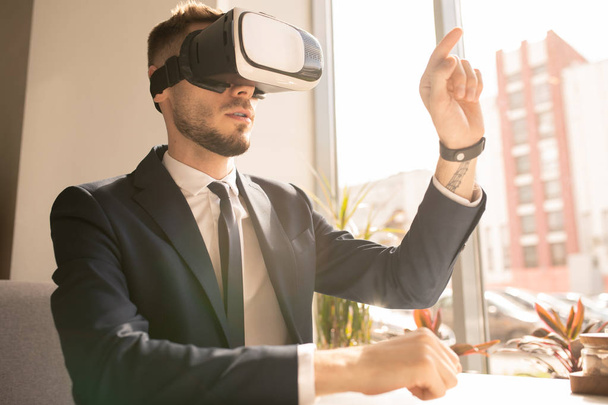 Young confident businessman in vr goggle pointing at virtual display while making presentation by table in cafe - Photo, image