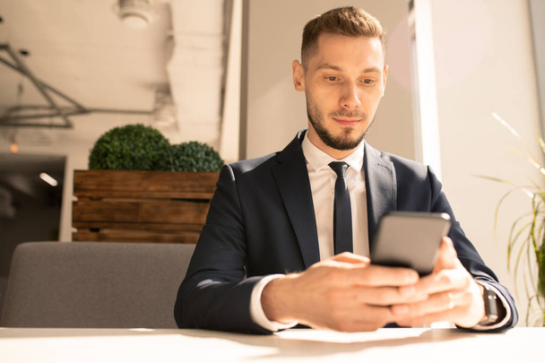 Молодий елегантний бізнесмен у костюмі і краватці, дивлячись повідомлення в смартфоні, обідаючи в кафе
 - Фото, зображення