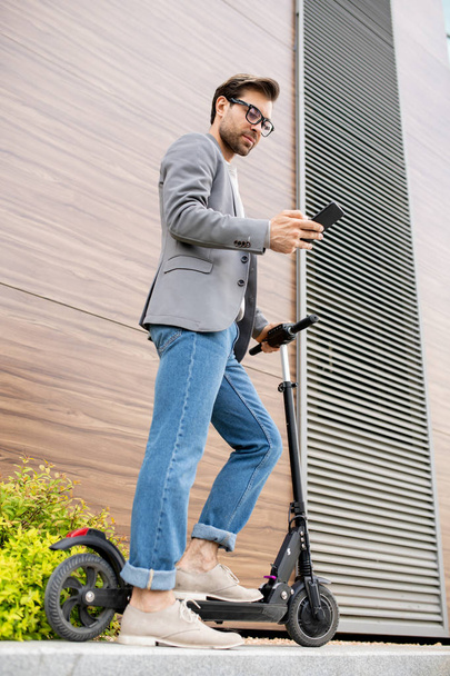 Joven móvil en smart casual parado junto a la pared de edificio moderno en su scooter y desplazándose en smartphone
 - Foto, imagen