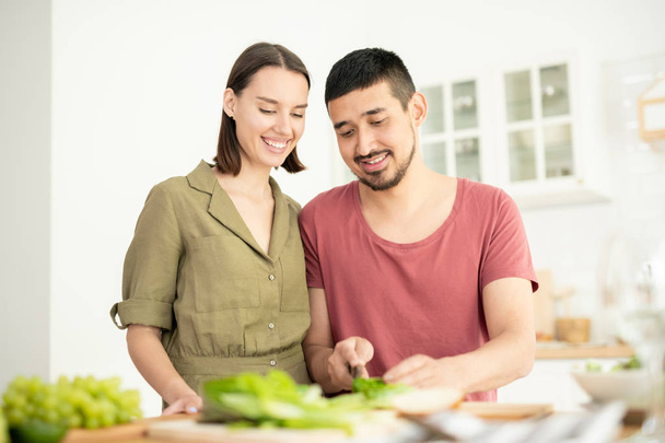Young handsome man spreading cheese or pate on slices of wheat bread or cutting vegetables for salad with his wife near by - Photo, Image