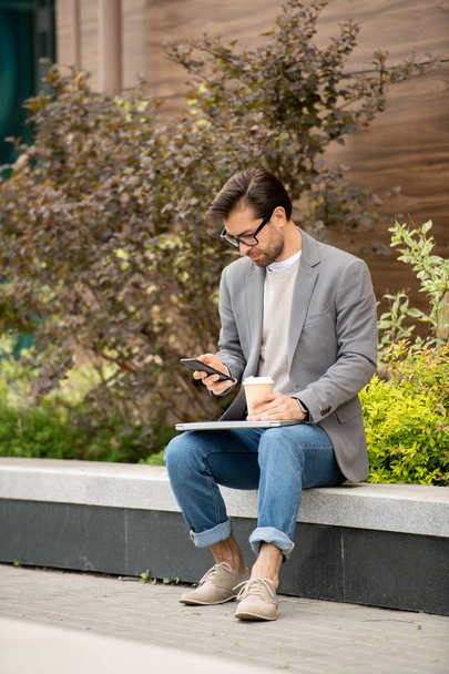 Kiireinen nuori johtaja smart casual älypuhelimella kun ottaa kahvia ulkona aamulla
 - Valokuva, kuva