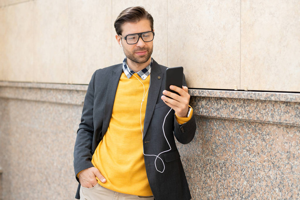 stylischer junger Mann in schickem Freizeitlook und Kopfhörer, der an einer Hauswand steht und Selfie macht, während er Musik hört - Foto, Bild