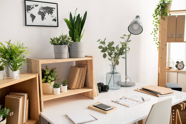Office desk with working supplies and picture of map on the wall over wooden shelves with flowerpots and books - Foto, imagen