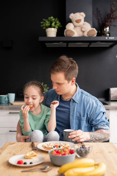 Contenida pequeña hija hambrienta sentada en las rodillas de los padres y comiendo crepe hecha por el padre cuidadoso, padre e hija desayunando juntos
 - Foto, imagen
