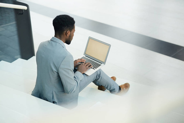 Joven hombre de negocios en elegante traje gris sentado en la escalera con portátil en las rodillas y navegar en la red para los datos financieros
 - Foto, imagen